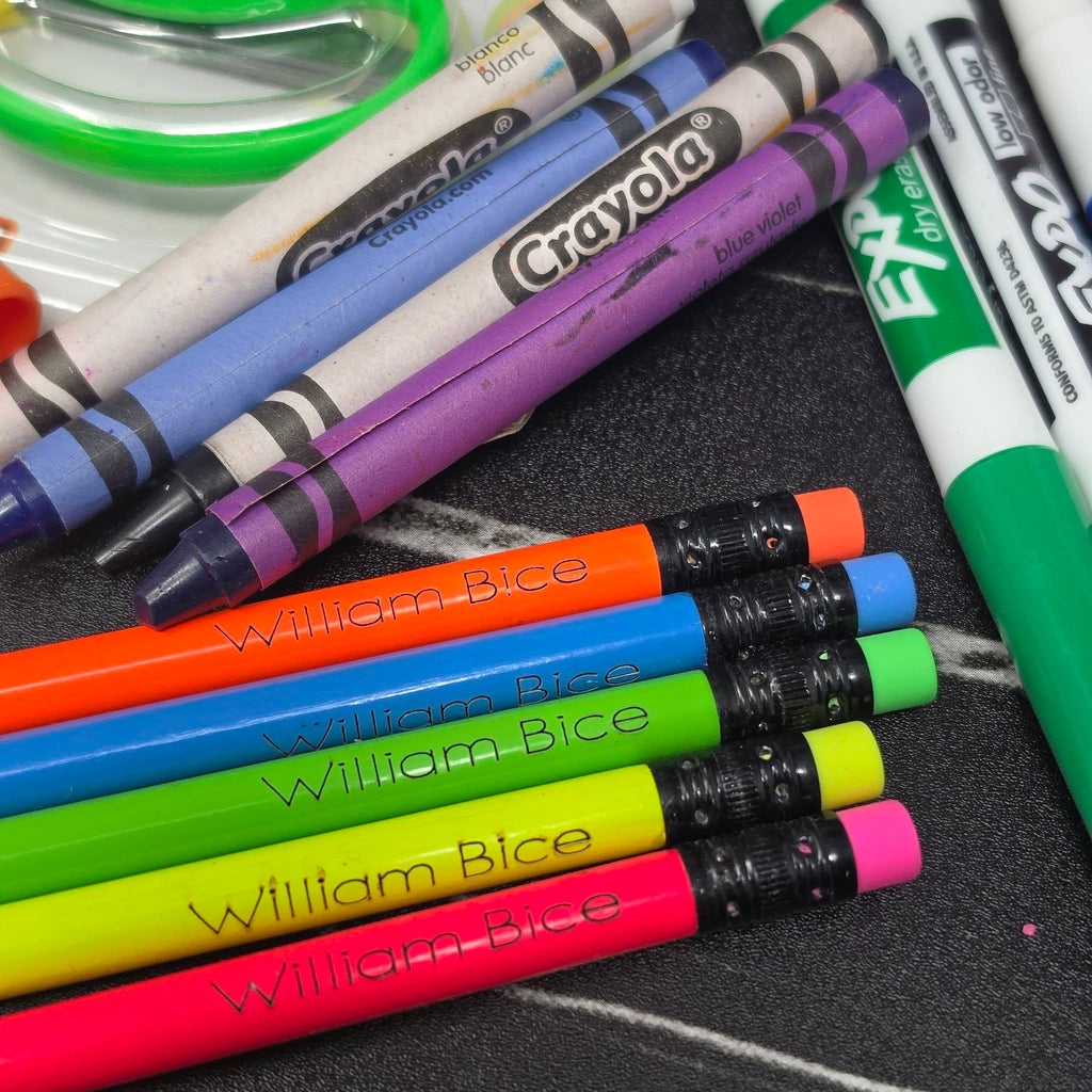A close-up arrangement of Crayola crayons and markers on a dark textured surface, with several pencils branded William Bice lying diagonally across the foreground.