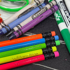 A close-up arrangement of Crayola crayons and markers on a dark textured surface, with several pencils branded William Bice lying diagonally across the foreground.