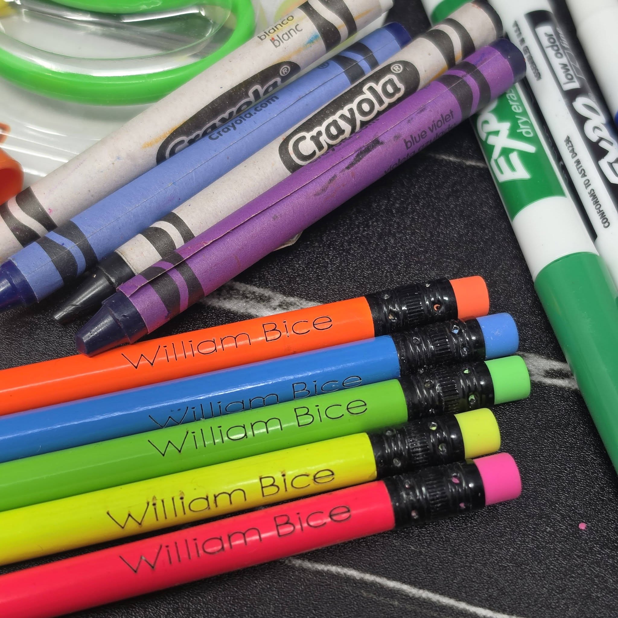 A close-up arrangement of Crayola crayons and markers on a dark textured surface, with several pencils branded William Bice lying diagonally across the foreground.